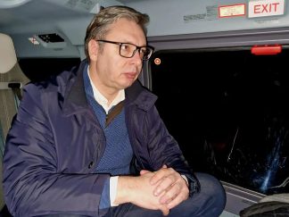 Predsenik Stbije Aleksandar Vučić sedi zamišljen u kaputu verovatno u helikopteru. Pored prozora vidi se znak Exit