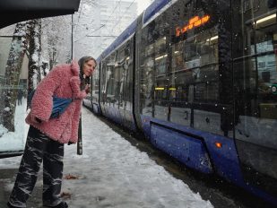 Žena gleda sa stanice tramvaja, tramvaj prolazi, sneg