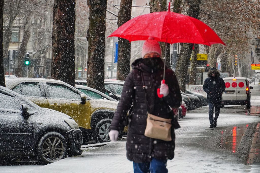 Žena sa kišobranom hoda ulicom dok pada sneg.