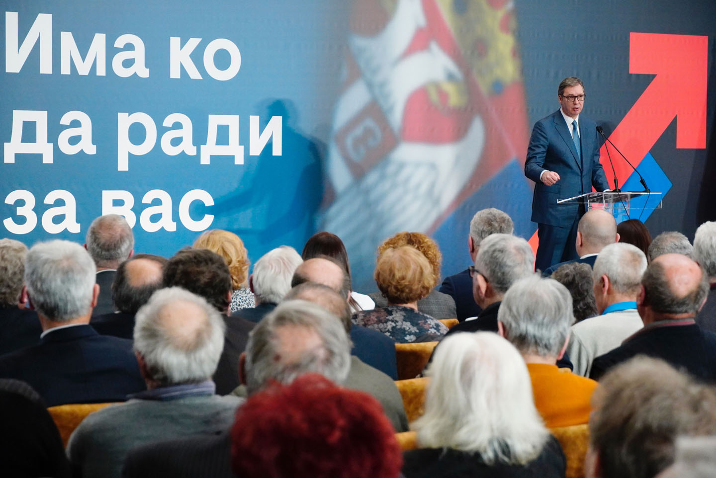 Predsednik Srbije Aleksandar Vučić u sali punoj starijih ljudi slikanim s leđa. Na bini dominira natpis 