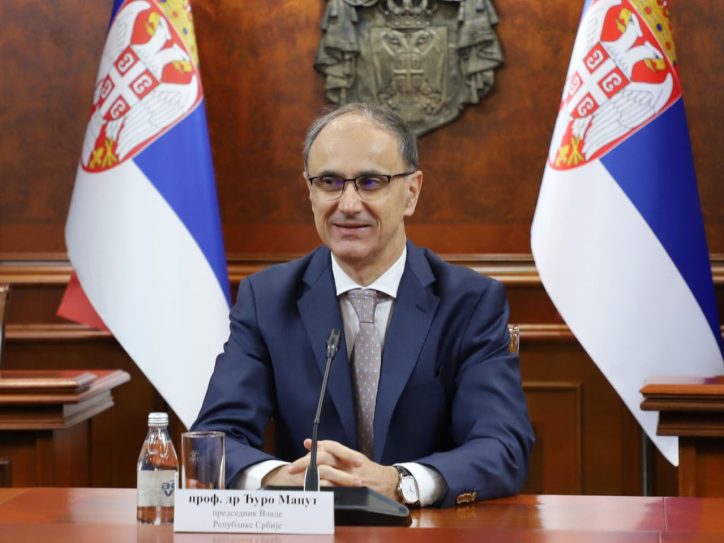 Premijer Đuro Macut između dve zastave sa grbom Srbije u teget odelu sa kravatom