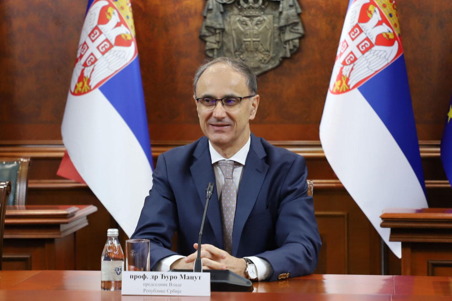 Premijer Đuro Macut između dve zastave sa grbom Srbije u teget odelu sa kravatom