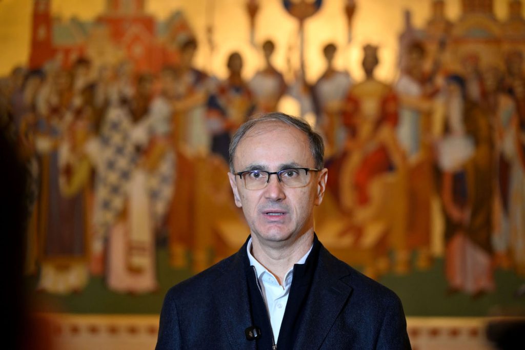 Premijer Srbije Đuro Macut sa tamnim sakoom i belom košuljom vez kravte i velikim pravoslavnim motivom u pozadini