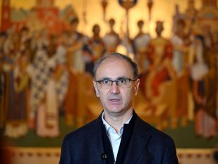 Premijer Srbije Đuro Macut sa tamnim sakoom i belom košuljom vez kravte i velikim pravoslavnim motivom u pozadini