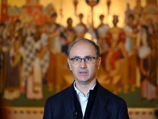 Premijer Srbije Đuro Macut sa tamnim sakoom i belom košuljom vez kravte i velikim pravoslavnim motivom u pozadini