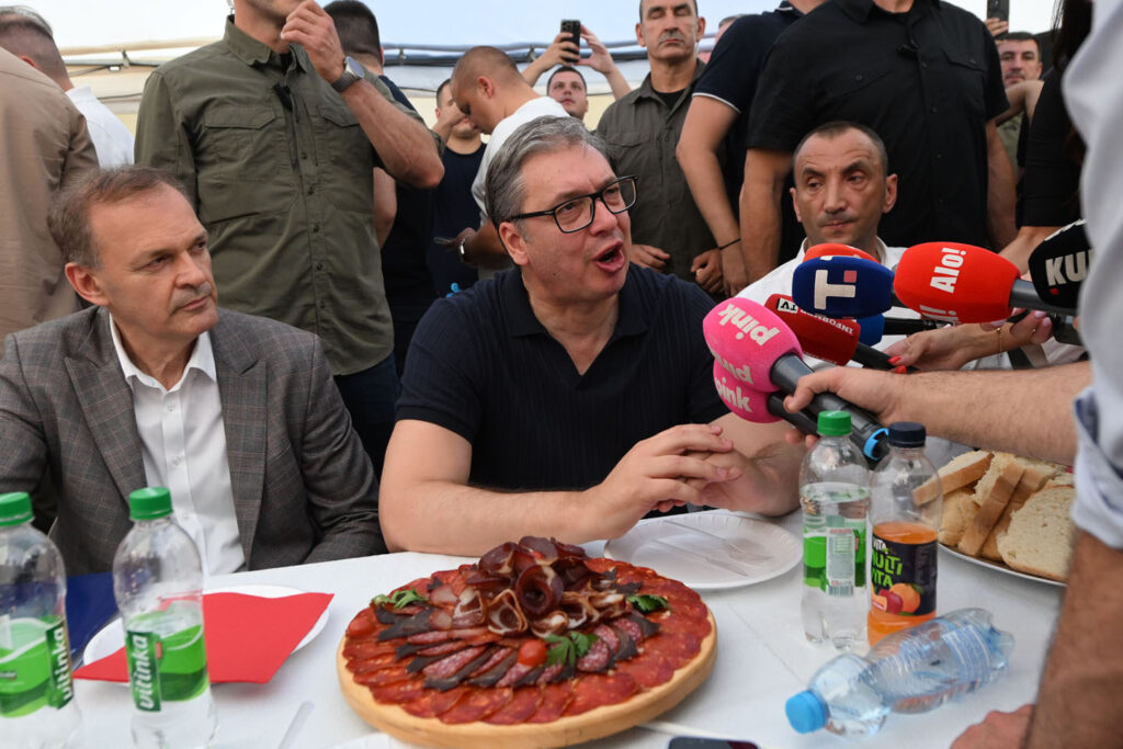 Aleksandar Vučić sedi za stolom pod šatorom, pred njim meze, sokovi i mikrofovni televizija